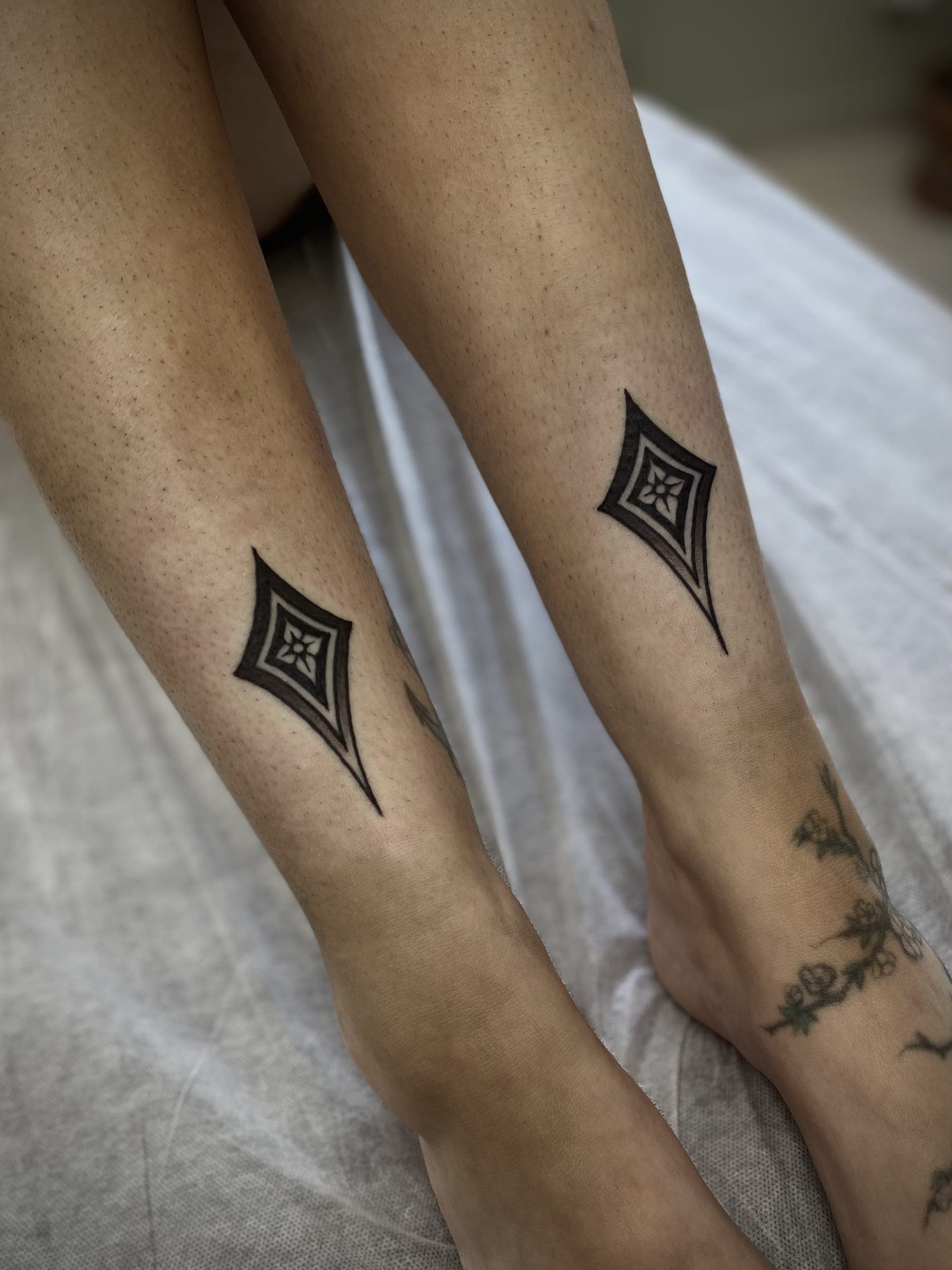 Two matching black diamond ornament tattoos on the lower legs of two people standing side by side