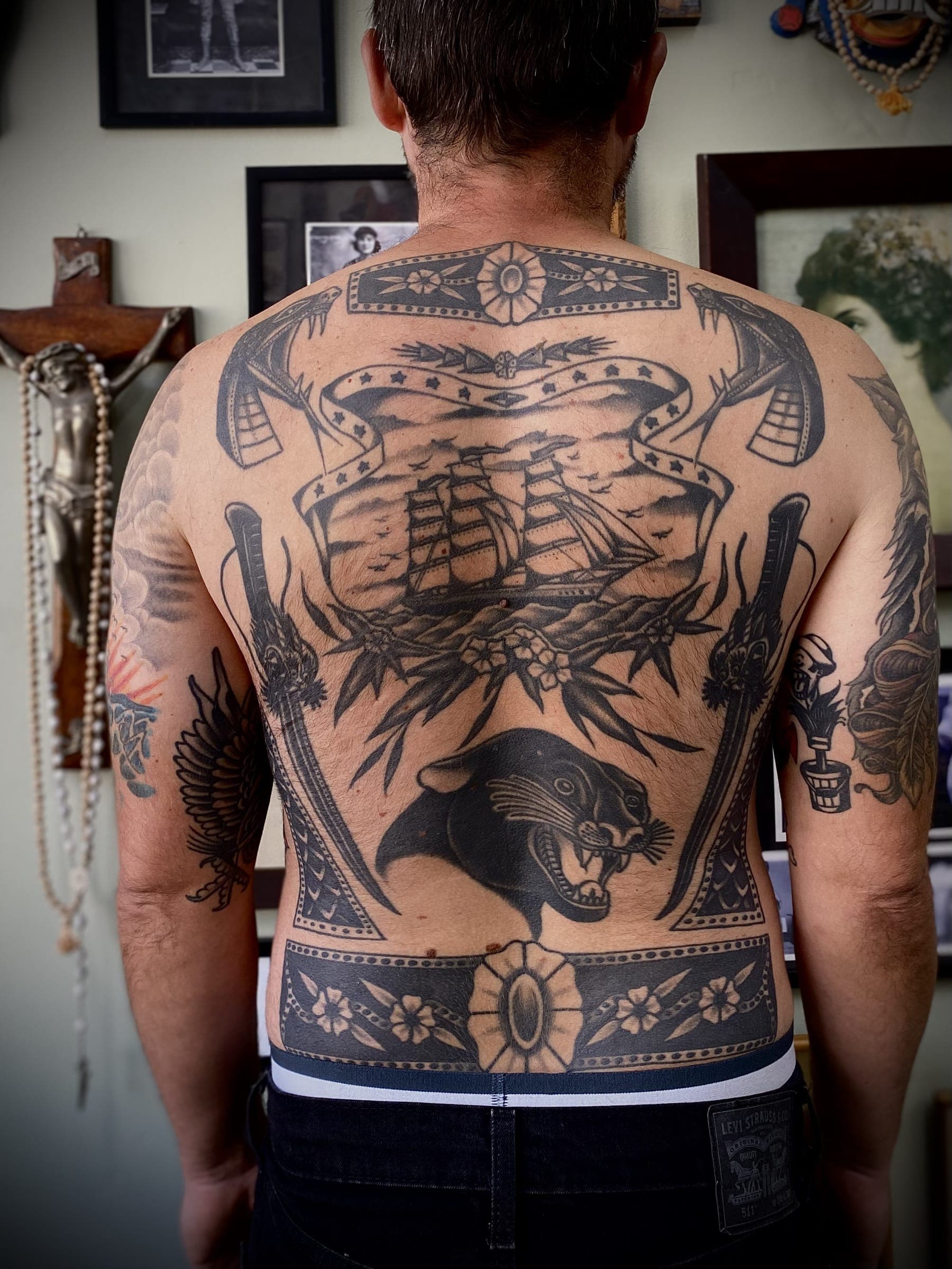 Full back tattoo with a tall ship banner above a snarling panther head framed by crossed daggers