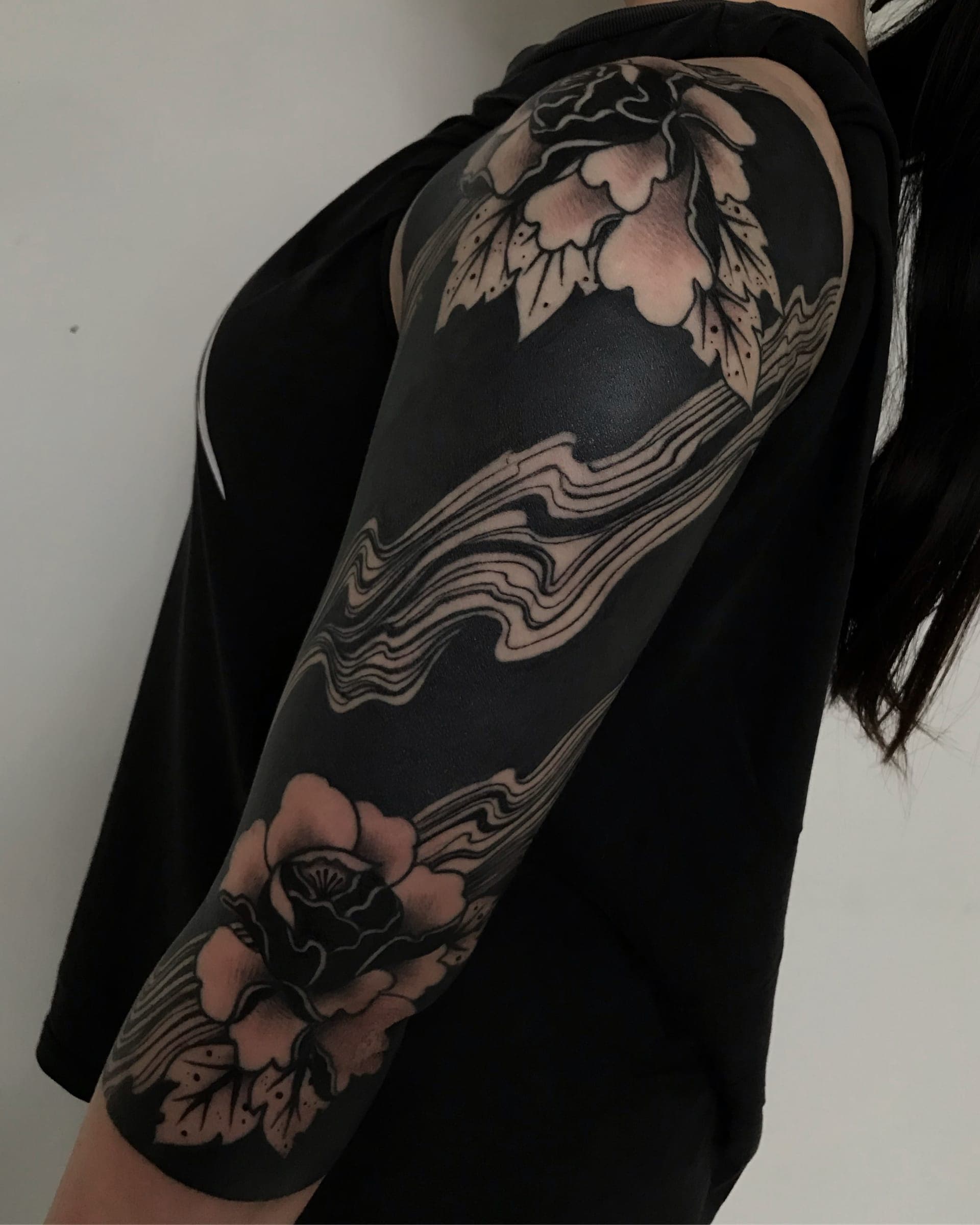 Blackwork upper arm with negative-space peony flowers and flowing water on a solid black background