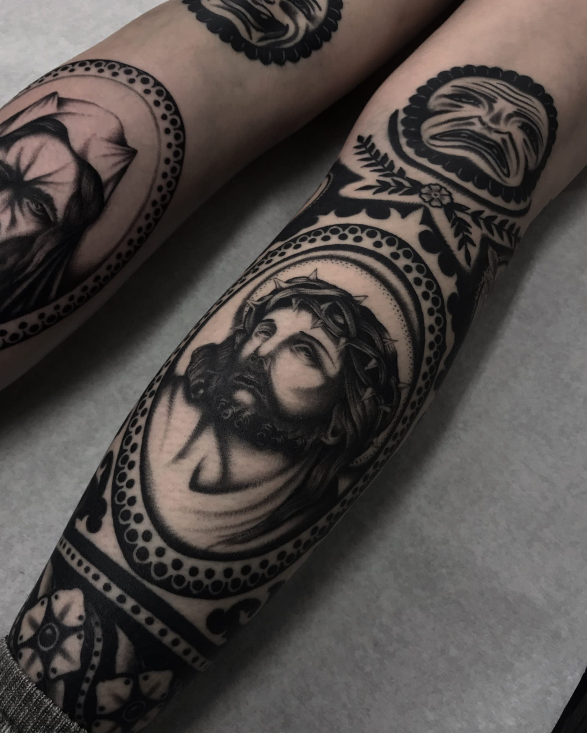 Black-and-grey crowned Christ portrait framed by ornamental blackwork and a beaded halo across the knee